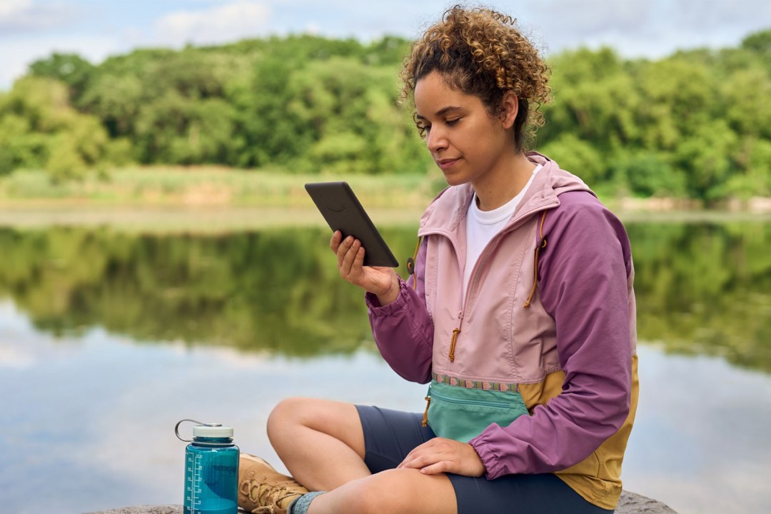 A woman sits by a lake reading on a Kindle e-reader