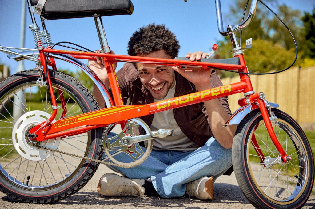 A man in jeans and a brown shirt sits on the floor and peers through the frame of a red Raleigh Chopper